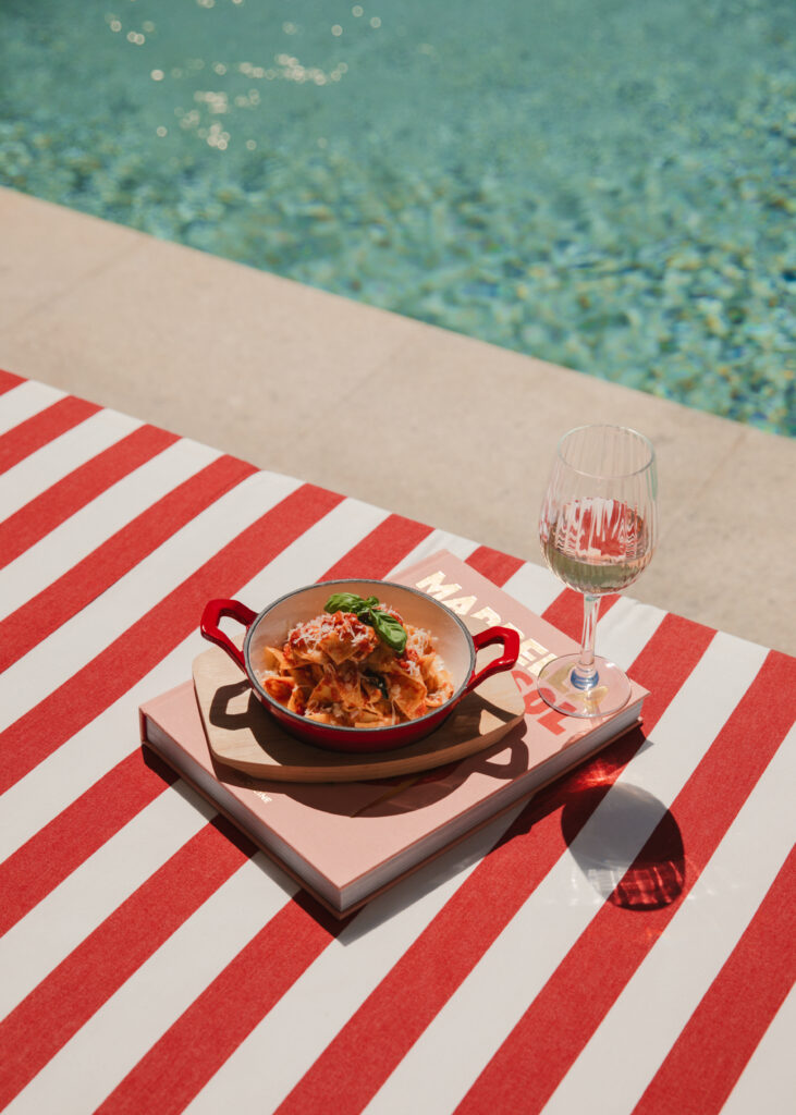 Plato de pasta y copa de vino sobre mesa a rayas junto a la piscina.